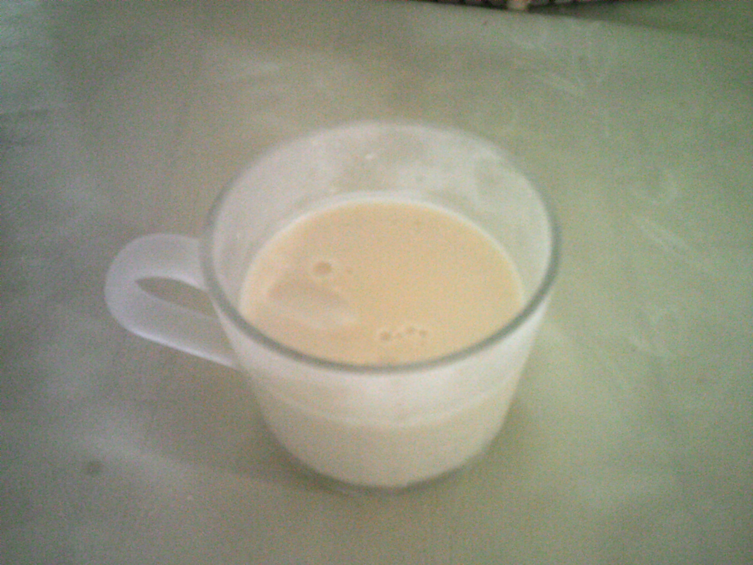 A cup containing lemon posset on a green background, a few bubbles can be seen in the mixture.