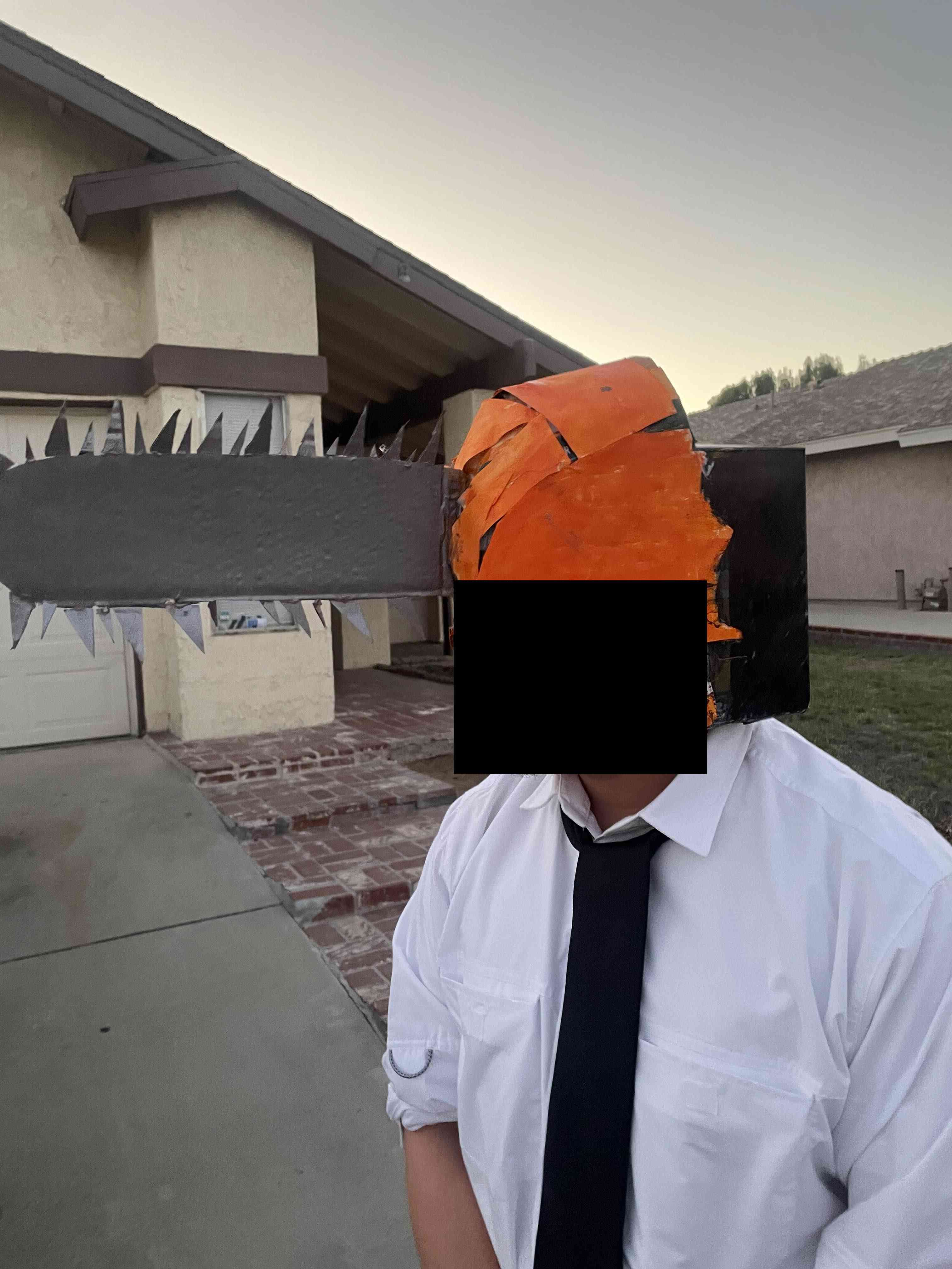 A short person standing in front of a house with a crude, orange, chainsaw shaped mask