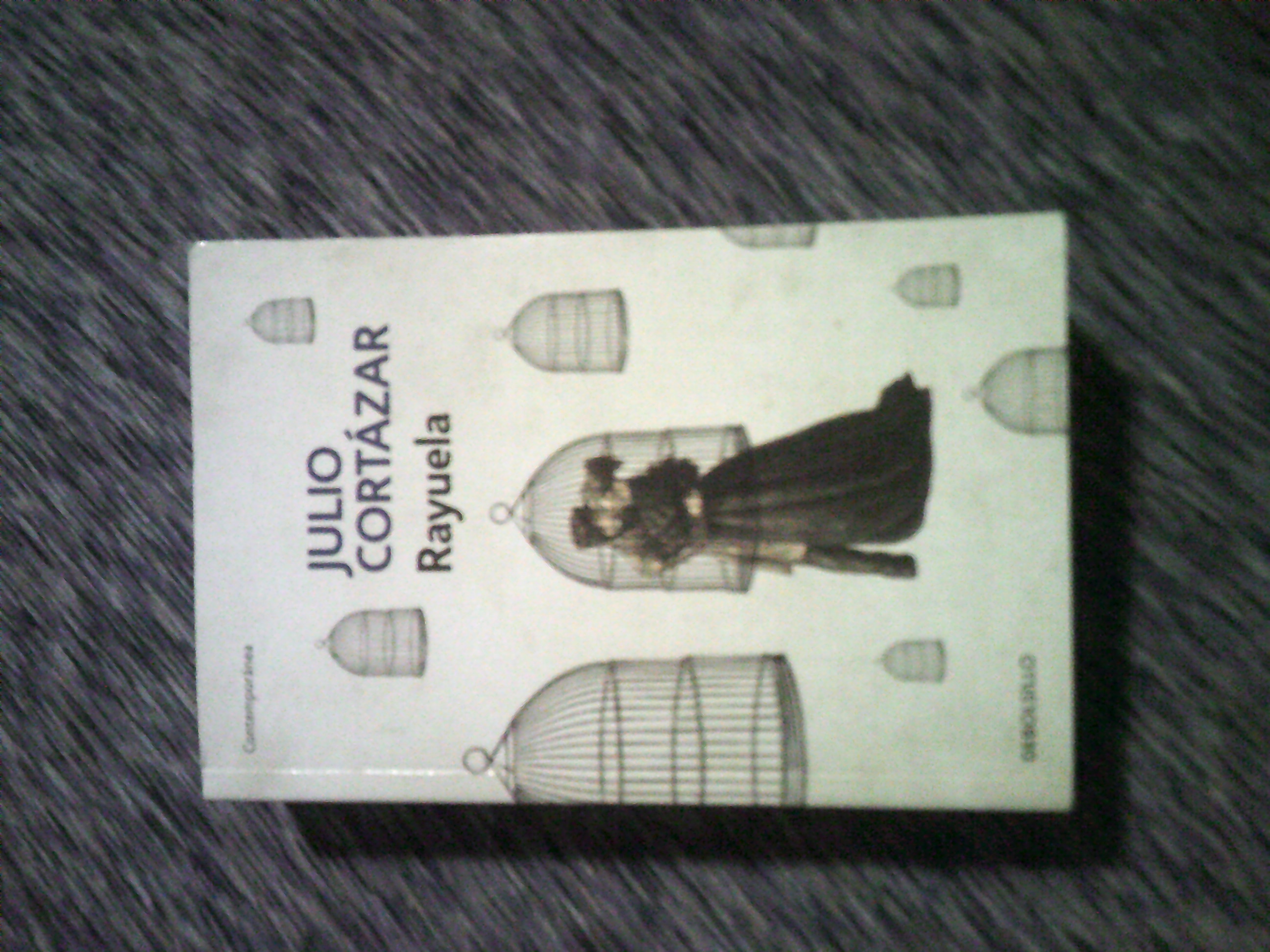 A small book titled 'Rayuela' written by Julio Cortázar. The cover depicts a couple dressed in dull victorian-era clothes kissing inside of a birdcage.